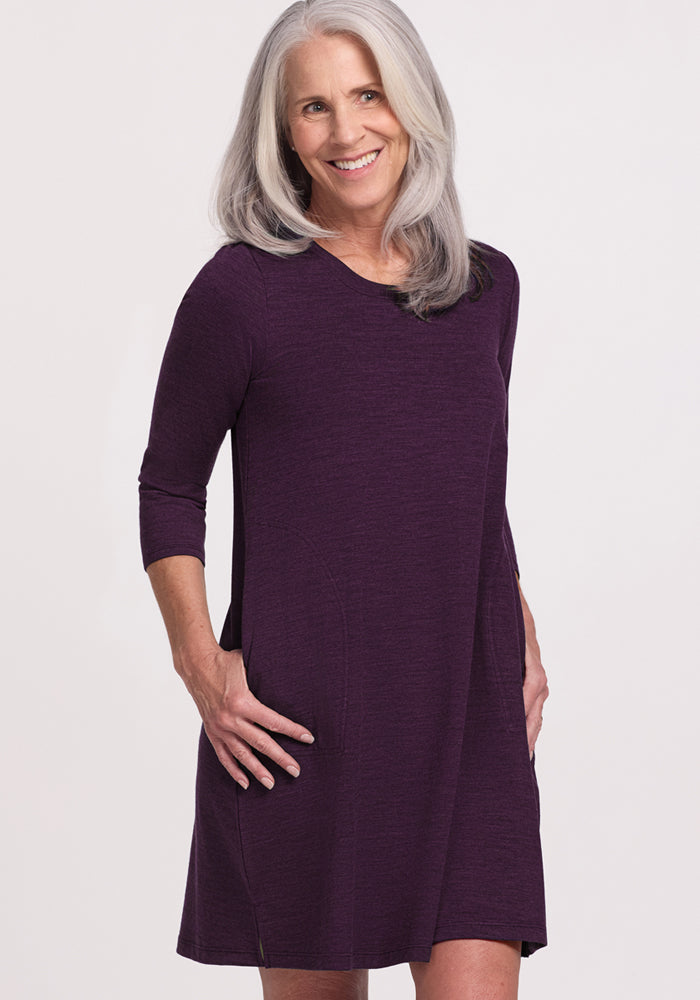 A smiling woman with long gray hair wears the Woolx Rissa A Line Dress in Deep Plum, a knee-length, long-sleeved Merino wool style with pockets, standing against a plain background—ideal for fall and winter layering.