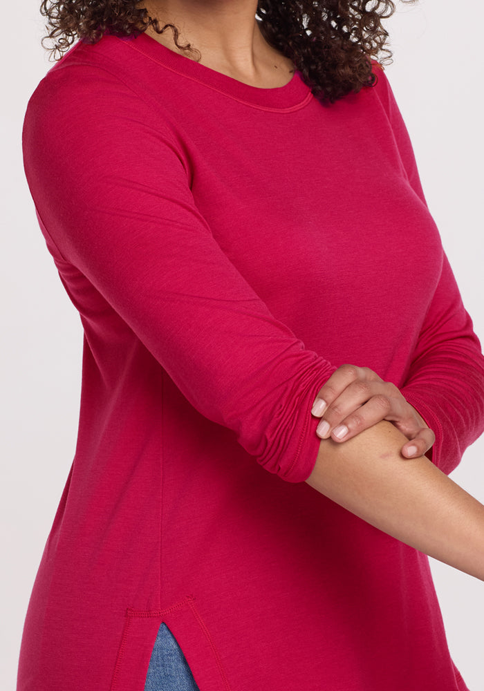 A person with curly hair models the Woolx Hazel Tunic in Poppy Spark, a long-sleeved bright pink merino wool top, pushing up one sleeve against a plain light background. Only their upper body is visible.