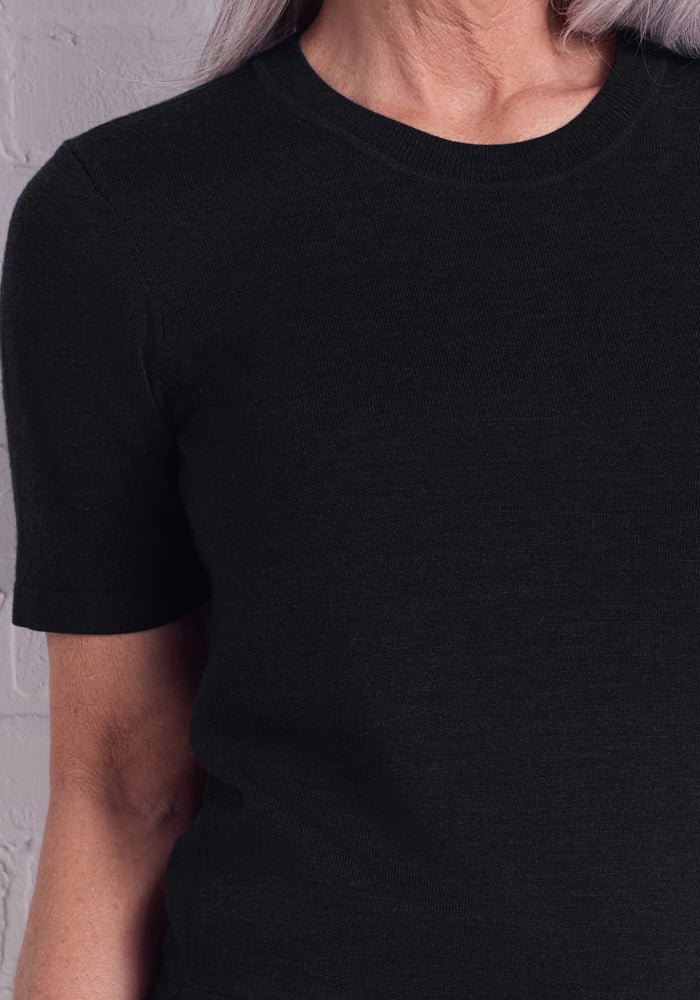 A person with light skin and gray hair models the Woolx Anna Crew Sweater in black, featuring short sleeves and a crew neck, shown from the upper torso up against a light brick wall.