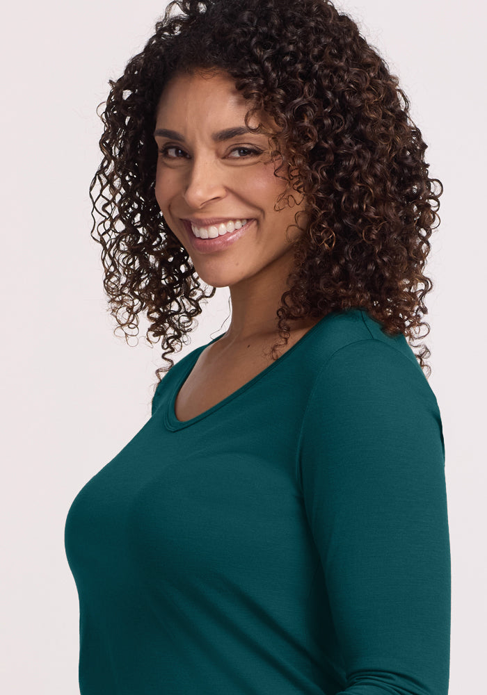A woman with curly dark hair smiles, looking sideways at the camera. She wears the Woolx Layla V Neck Top in Riverbank Green, a cozy long sleeve known for moisture-wicking, standing against a light, neutral background.