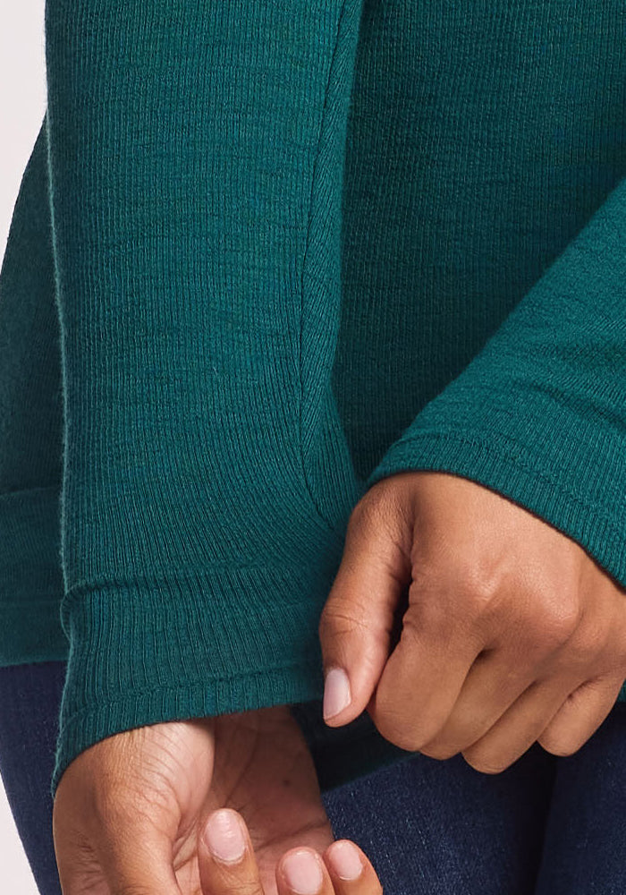 A close-up of a person wearing the Woolx Reese Ribbed Henley in Riverbank Green with dark jeans, gently pulling at the sleeve of the other arm.