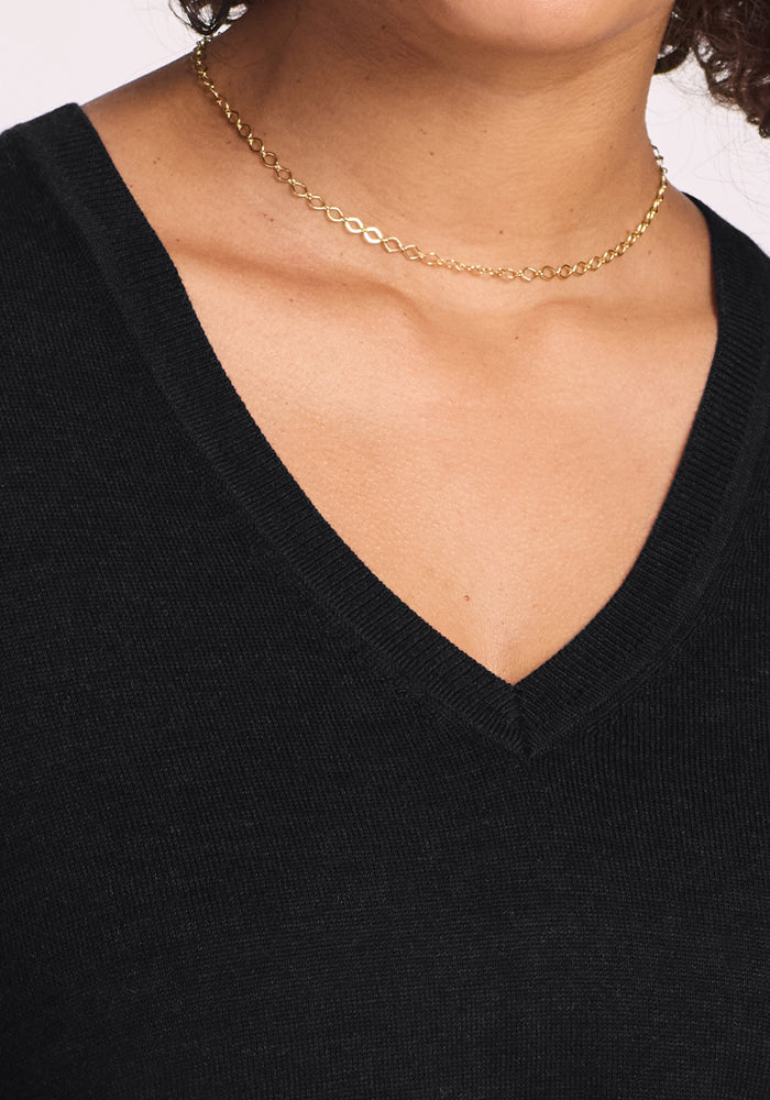 Close-up of a person wearing the Woolx Francesca Sweater in black, featuring breathable Merino wool and a V-neck. A delicate gold chain necklace accents the neckline, while curly hair frames the neck, collarbone, and upper chest.