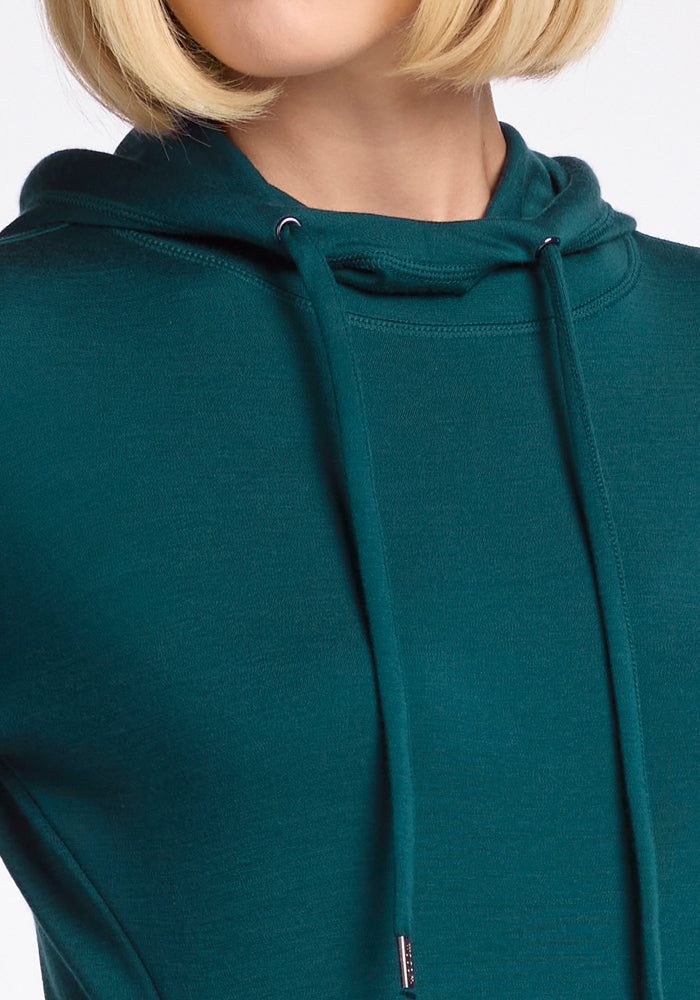 A close-up of a person in the Woolx Callie Hoodie in Riverbank Green, featuring ultra-soft fabric for cozy cold-weather layering. The individual’s short blonde hair and partially out-of-frame face add visual interest.