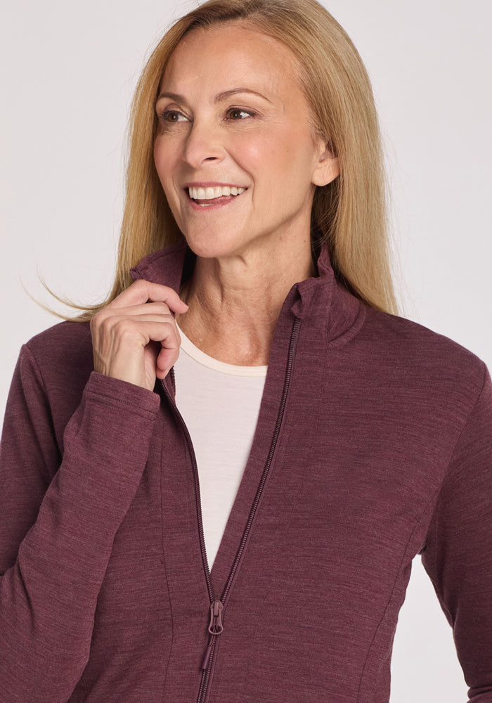 A smiling woman with long blonde hair wears the Woolx Finley Zip Up Sweatshirt in Grape Harvest Melange over a white top, holding the collar and highlighting its odor-resistant comfort against a light background.