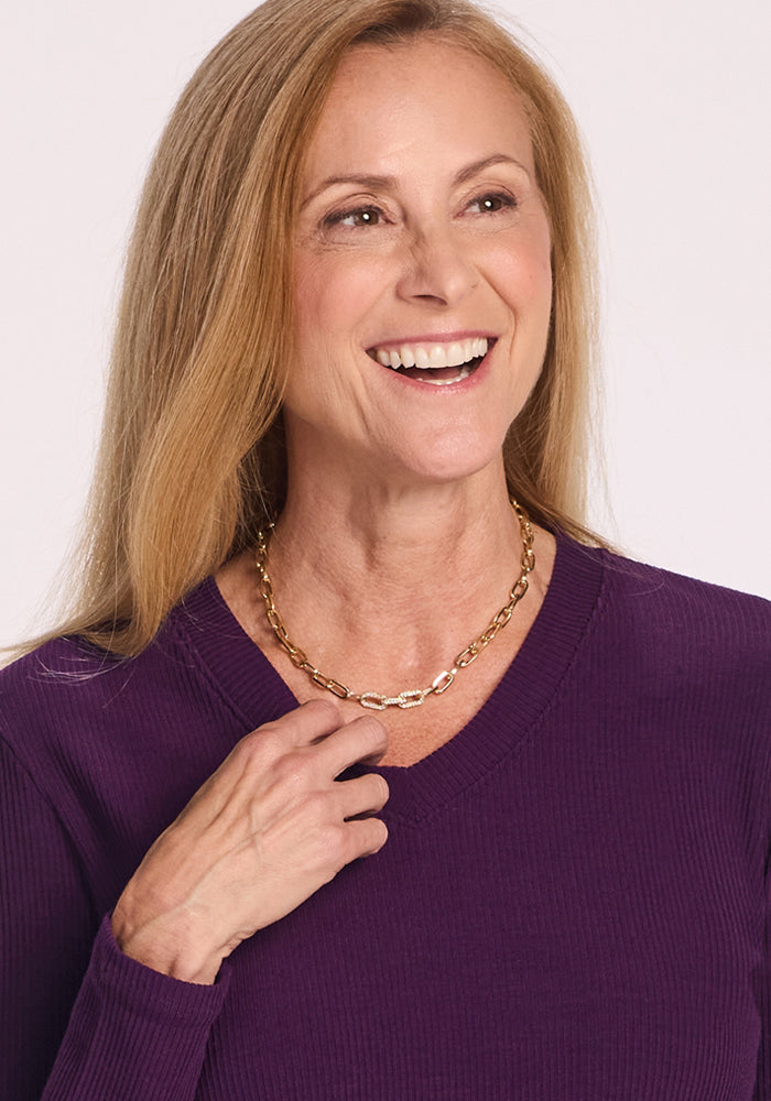 Smiling woman with long blonde hair wears the Woolx Evie Ribbed Tunic in Majestic Purple, a cozy V-neck women’s top, styled with a gold chain necklace.