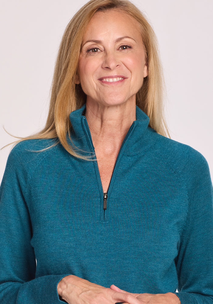 A woman with long blonde hair, wearing the Woolx Collins Half Zip Sweater in Harbor Teal, smiles gently against a plain light background.