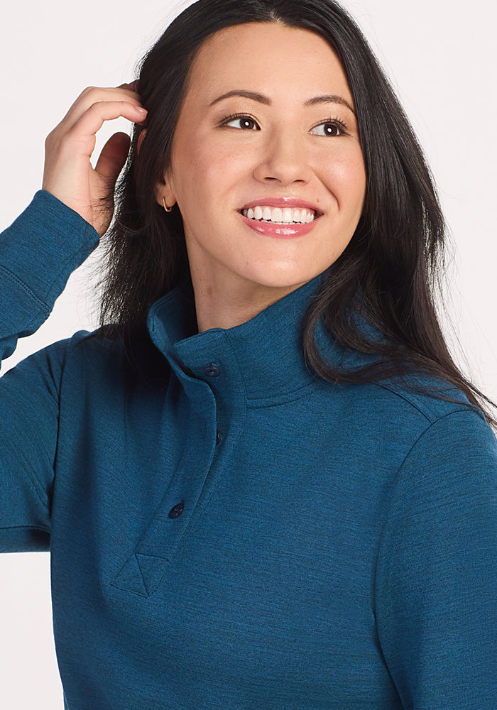 A smiling woman with long dark hair, wearing the Woolx Annie 3 Button Pullover in Majolica Blue, poses with one hand touching her hair against a light background.