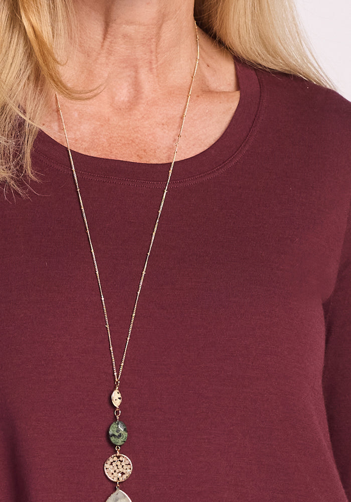 A woman wearing the Woolx Rissa A Line Dress in Toasted Garnet and a long gold necklace with green and circular pendants. The image is cropped to her neck, shoulders, and upper chest—ideal for fall and winter layering.