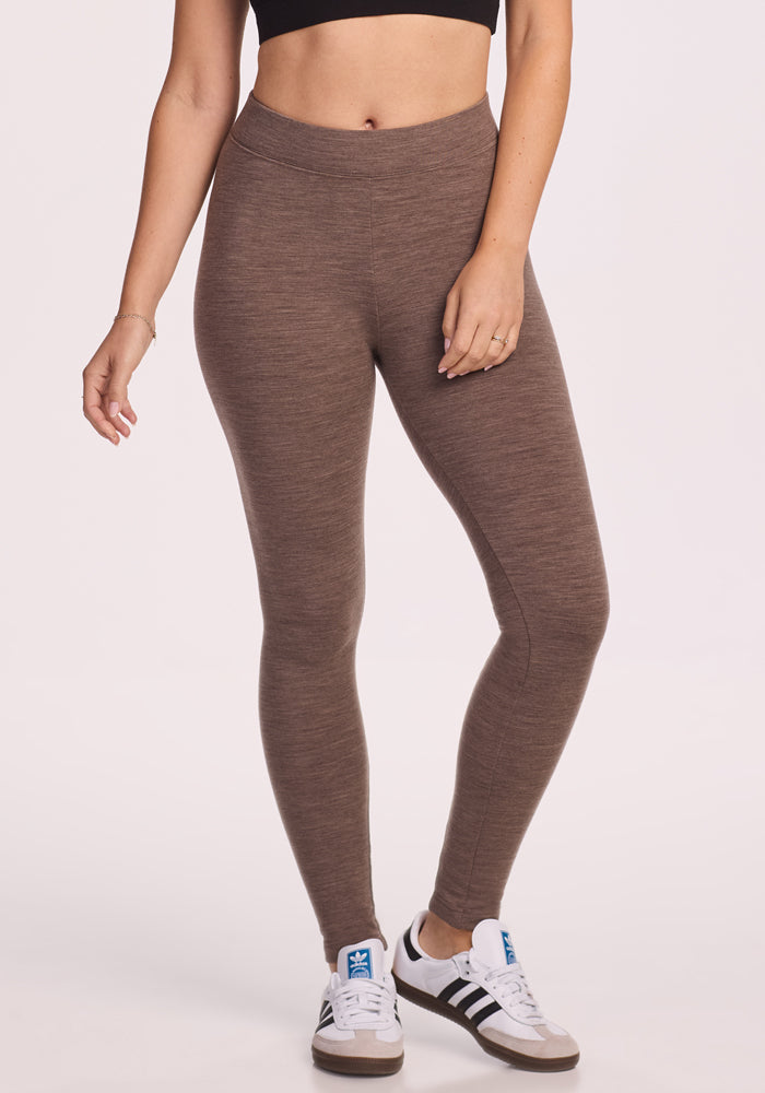 A person wears Woolx Stella Leggings in Simply Taupe, a black crop top, and white sneakers with black stripes, standing against a plain light background.