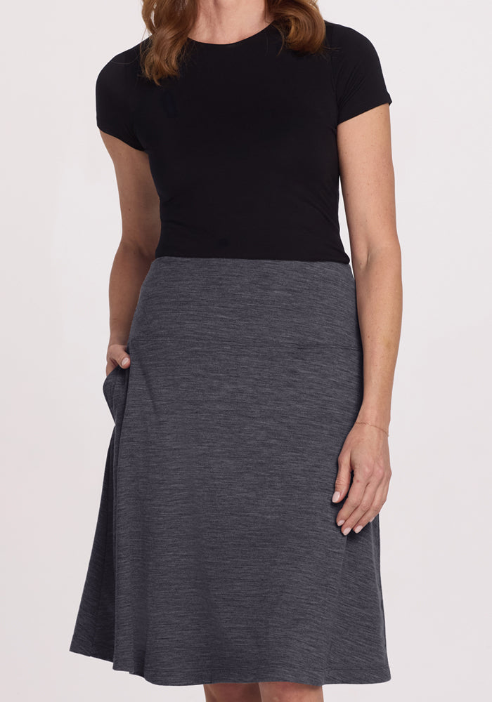 A woman wears a short-sleeved black top and the Woolx Marilyn Flowy Midi Skirt in Cinder Melange, knee-length with her right hand in a pocket, against a plain, light background.