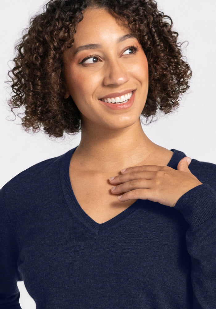 A woman with curly brown hair smiles and looks to the side, wearing the Woolx Francesca Sweater in Storm Cloud, her hand near her collarbone. The background is light and plain.