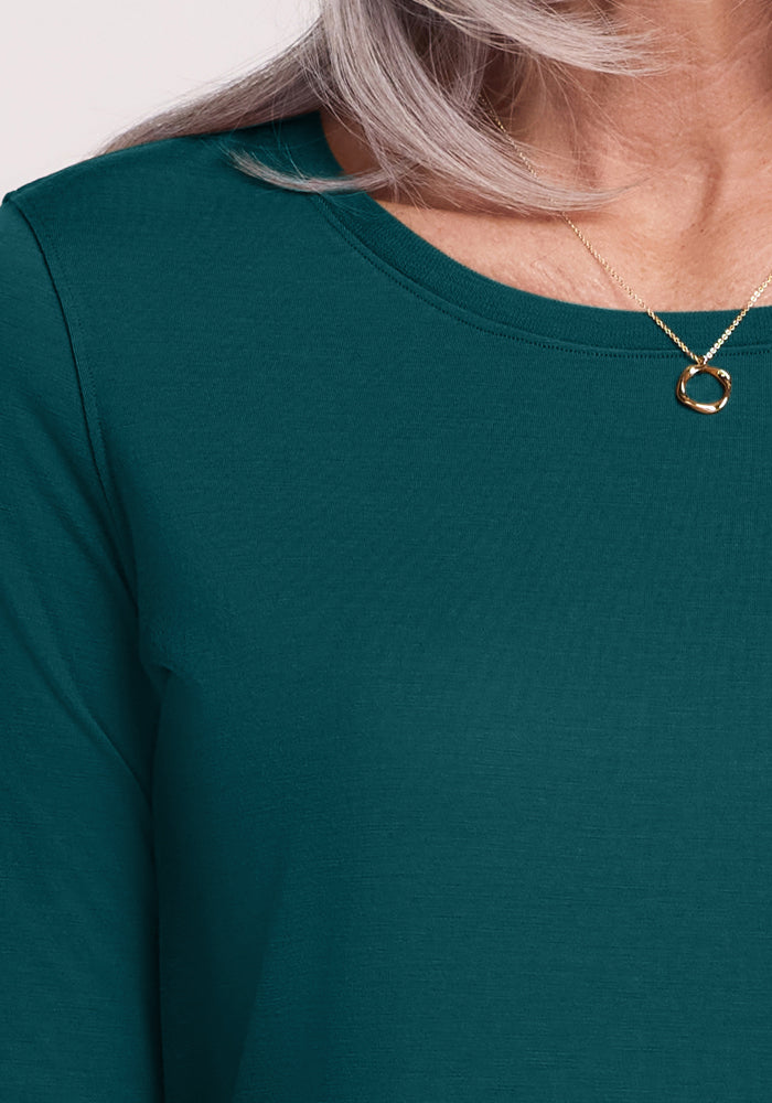 A close-up of a person wearing the Woolx Jenny 3/4 Sleeve Crew Neck in Riverbank Green and a gold circular pendant necklace. Only their long gray hair, upper chest, part of the shoulder, and neck are visible.