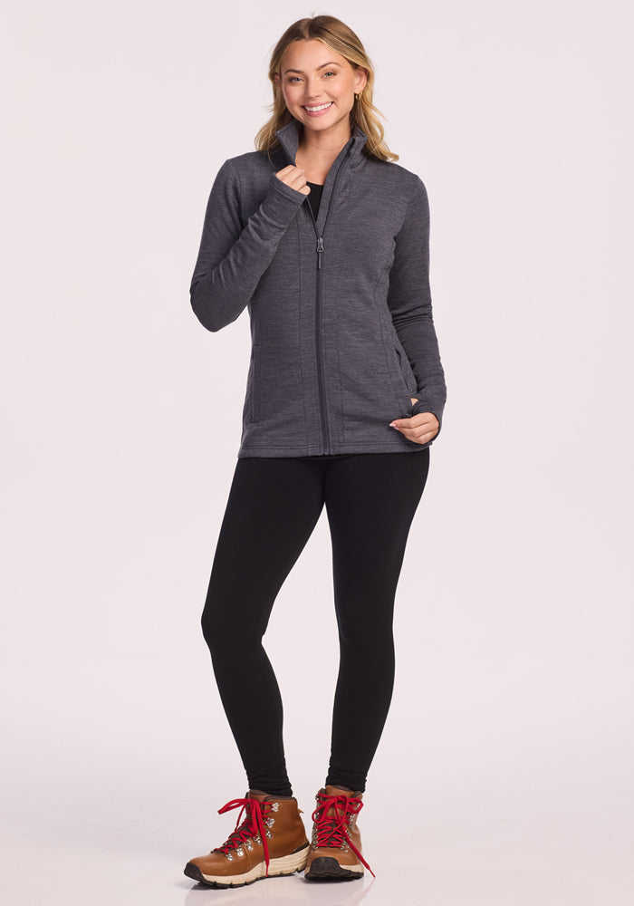 A woman smiles, wearing the Woolx Finley Zip Up Sweatshirt in Cinder Melange, black leggings, and brown hiking boots with red laces. She has one hand in her pocket and the other on her collar, posing against a plain light background.