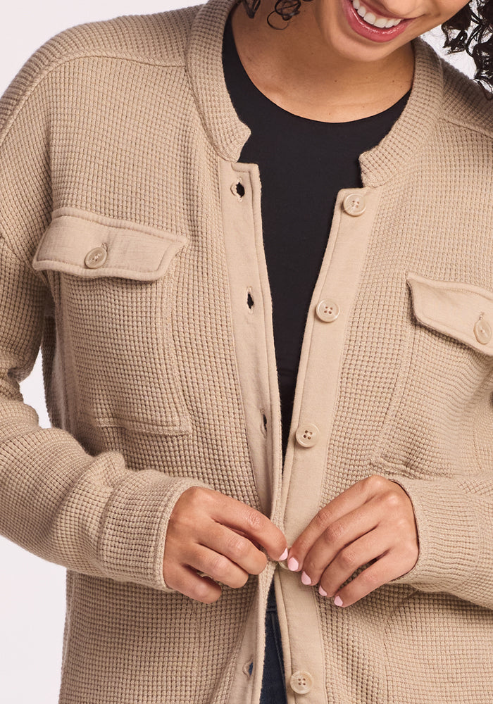 A woman smiles while buttoning the Woolx Oaklynn Waffle Shacket in Biscotti over a black top. Only her torso and lower face are shown.