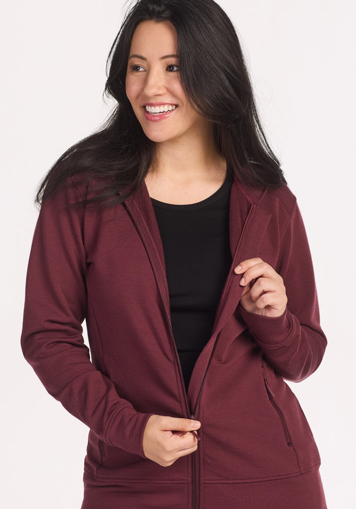 A woman with long dark hair wears the Woolx Darby Zip Up Hoodie in Toasted Garnet over a black top, smiling and looking to the side as she unzips her jacket against a plain light background.