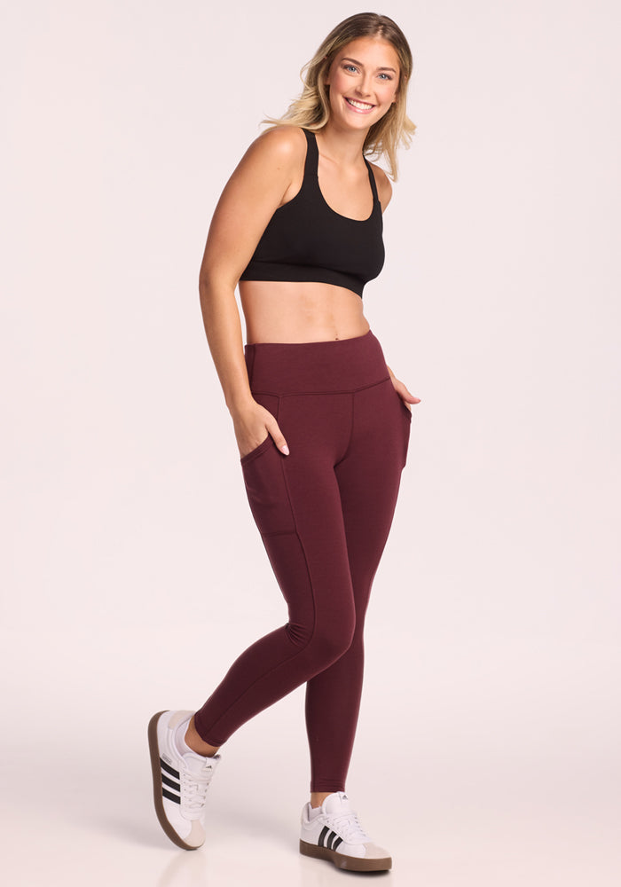 A smiling woman wearing a black sports bra, white Adidas sneakers, and Woolx Piper Pocket Petite leggings in Toasted Garnet stands posing against a plain light background.