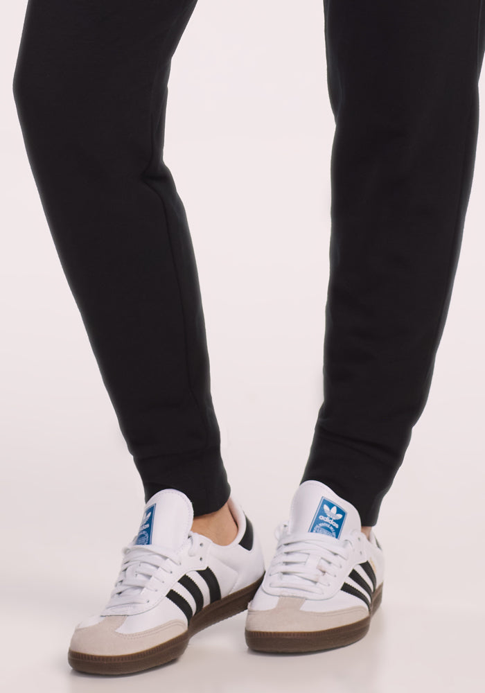 A person wearing Woolx Willa Joggers Petite in black and white Adidas sneakers with black stripes stands on a light-colored surface, showing only their lower legs and feet.