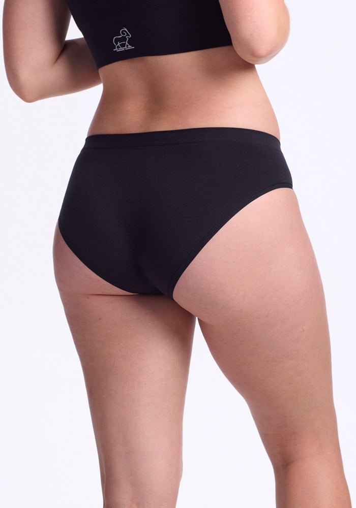 A person wearing the Woolx Maeve Bikini - Black, featuring high-waisted, moisture-wicking underwear and a matching black top, stands with their back to the camera against a plain white background.