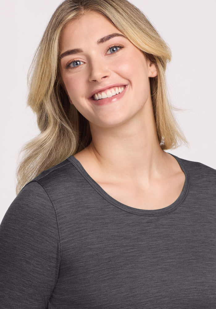 A young woman with long blonde hair smiles warmly while wearing the Woolx Remi Long Sleeve T-Shirt in Cinder Melange, posed against a light background.