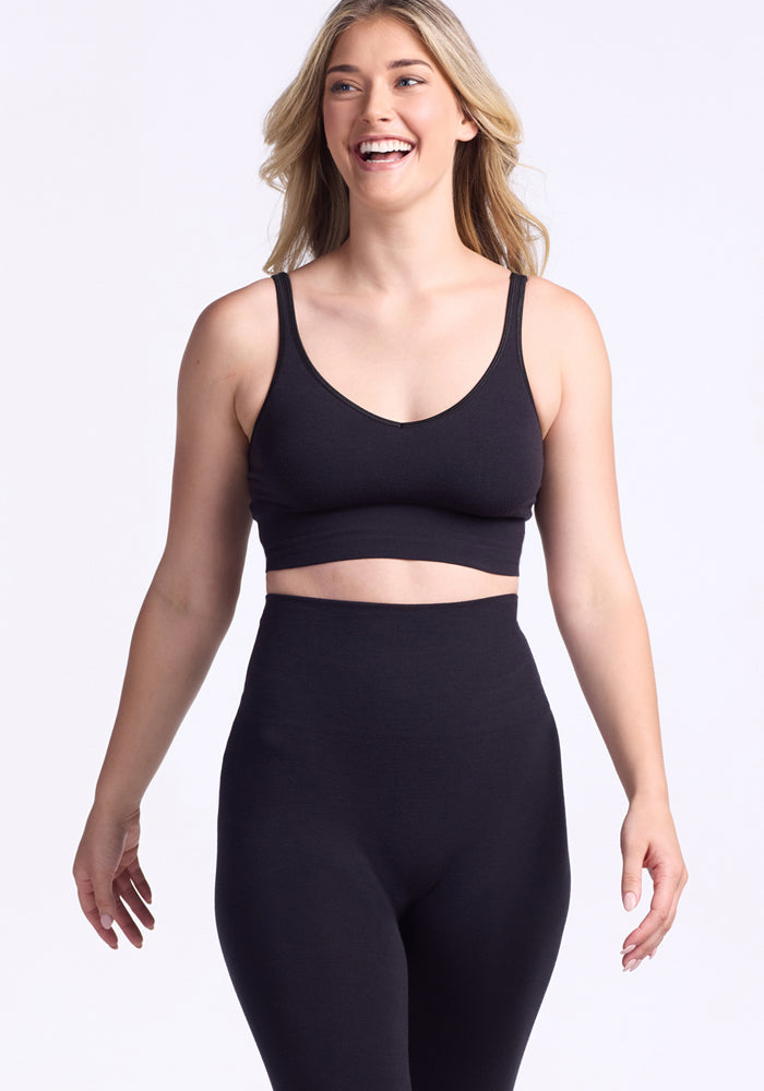 A woman with long blonde hair, wearing a black sports bra and Woolx Ellis Seamless Petite Tights in black, smiles while standing against a plain white background.