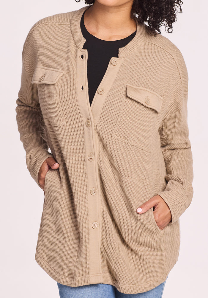 A person wears the Woolx Oaklynn Waffle Shacket in Biscotti, featuring chest flap pockets and textured knit, layered over a black top with light blue jeans. The warm shacket is perfect for layering; the person's face is partially out of frame.