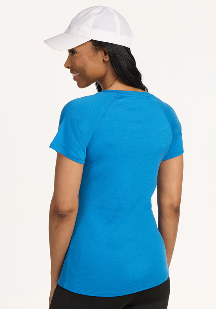 A woman wearing the Woolx Mia V Neck - Wave Rider stands facing away with her head slightly turned to the side. She also wears a white baseball cap against a plain, light background.