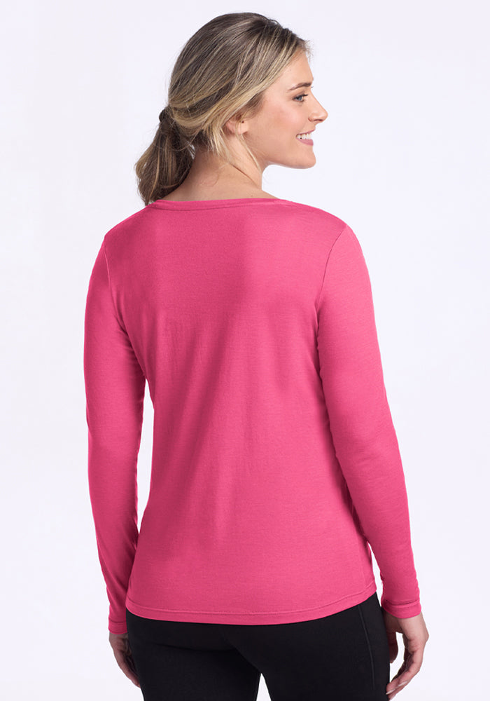 A woman with light brown hair in a low ponytail wears the Woolx Millie Long Sleeve in Raspberry Fizz and black pants. She stands with her back to the camera, smiling and looking left against a plain white background.