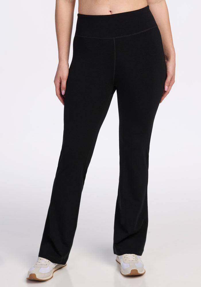 Wearing Woolx’s Morgan Flare Pants in black, a person stands against a plain white background, visible only from midriff to feet, paired with white sneakers. 