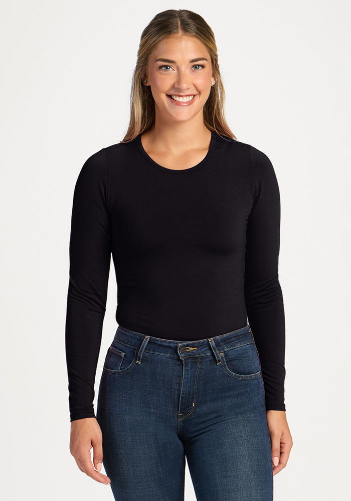 Woman smiles in Woolx Margot Bodysuit—black, long-sleeve merino wool—styled with blue jeans, white backdrop. 