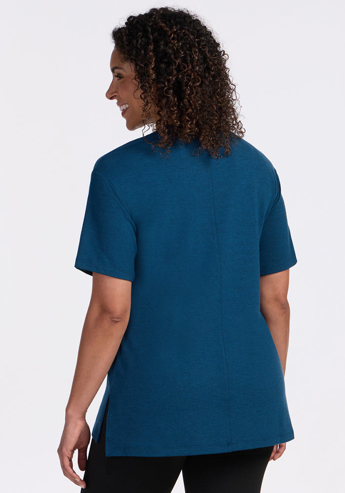 A woman with curly hair is seen from behind wearing the Woolx Audrey Tee in Majolica Blue and black pants, standing against a plain white background.