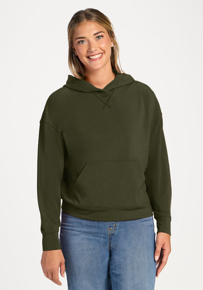 Smiling woman wears Woolx Lizzy Hoodie - Everleaf, paired with blue jeans, against a plain white background.