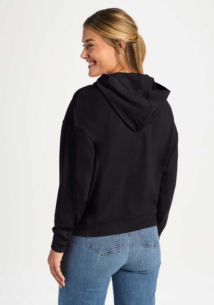 Woman smiling in the Woolx Lizzy Hoodie - Black and blue jeans, shown from the back on a plain background.