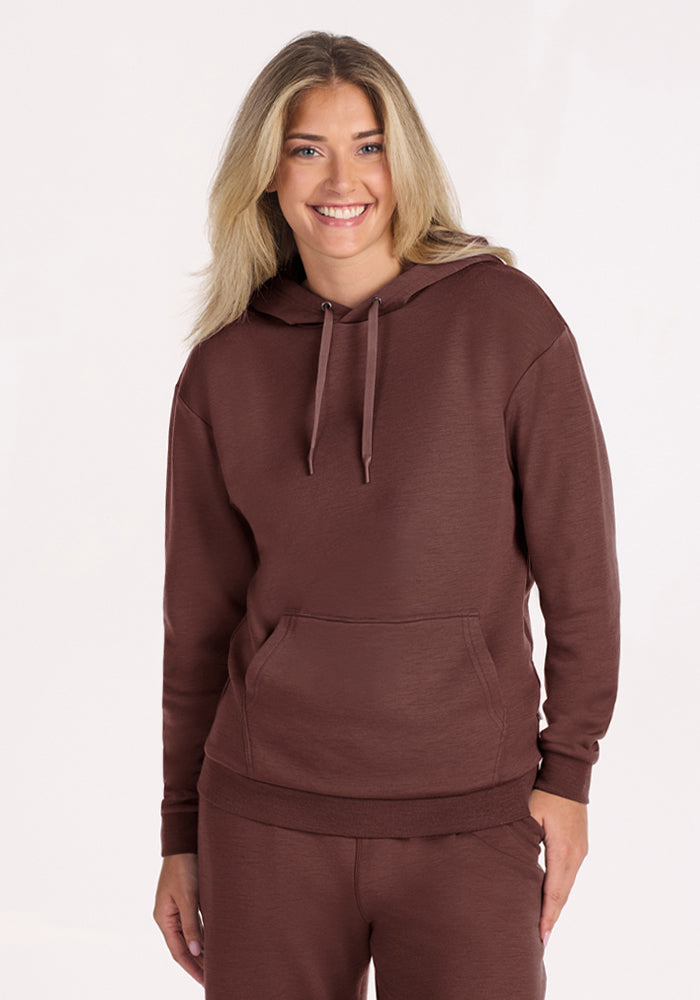 Smiling blonde in Woolx Avery Hoodie - Canyon Clay, with matching pants, stands against a plain light background. 