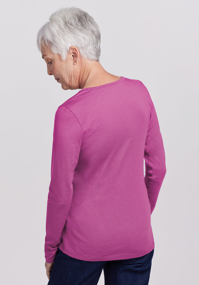 An older woman with short gray hair stands facing away, wearing the Woolx Remi Long Sleeve T-Shirt in Purple Wine and dark pants against a light background.