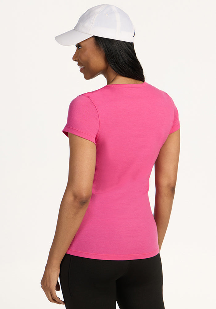 A woman faces away, wearing the Woolx Addie Short Sleeve Crew in Raspberry Fizz—an odor-free, short-sleeve shirt—with black pants and a white baseball cap against a plain light background.