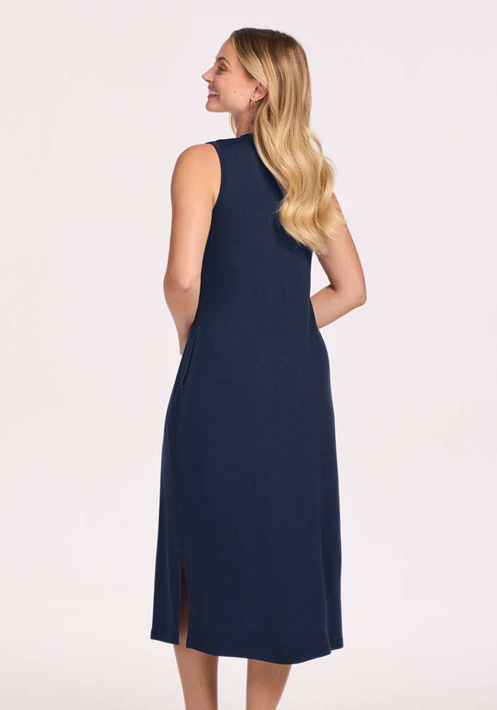 A woman with long blonde hair is wearing the Woolx Cassie Dress in Deep Navy, a sleeveless, midi-length Merino wool dress. She stands smiling with her hands behind her back—perfect as a wrinkle-resistant travel dress.