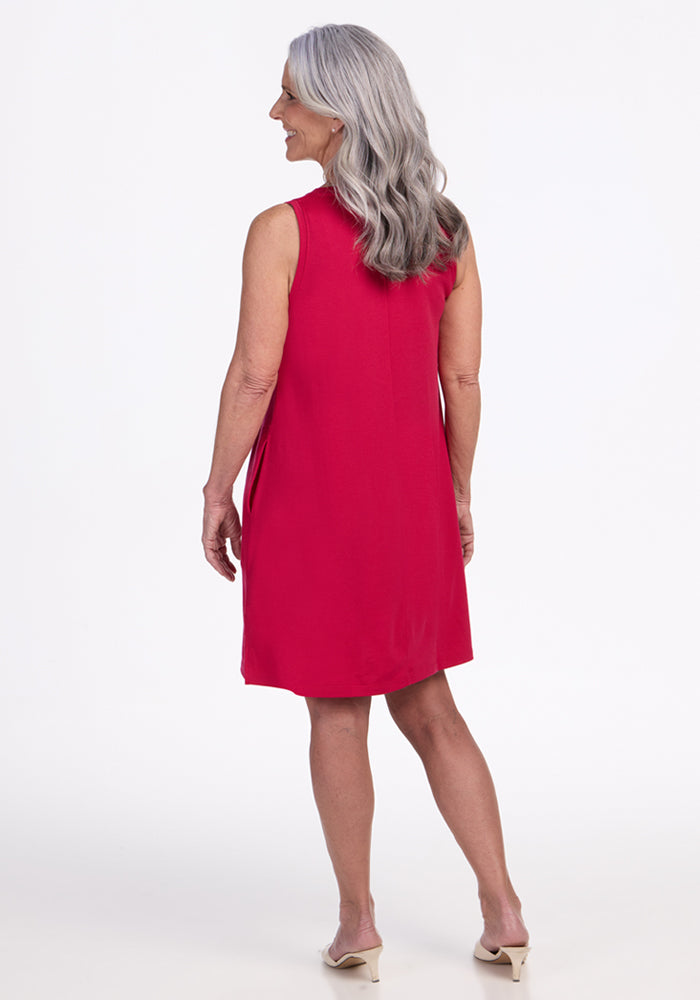 An older woman with long gray hair stands facing away, wearing the Woolx Clara Dress in Poppy Spark and beige heeled shoes against a plain white background.