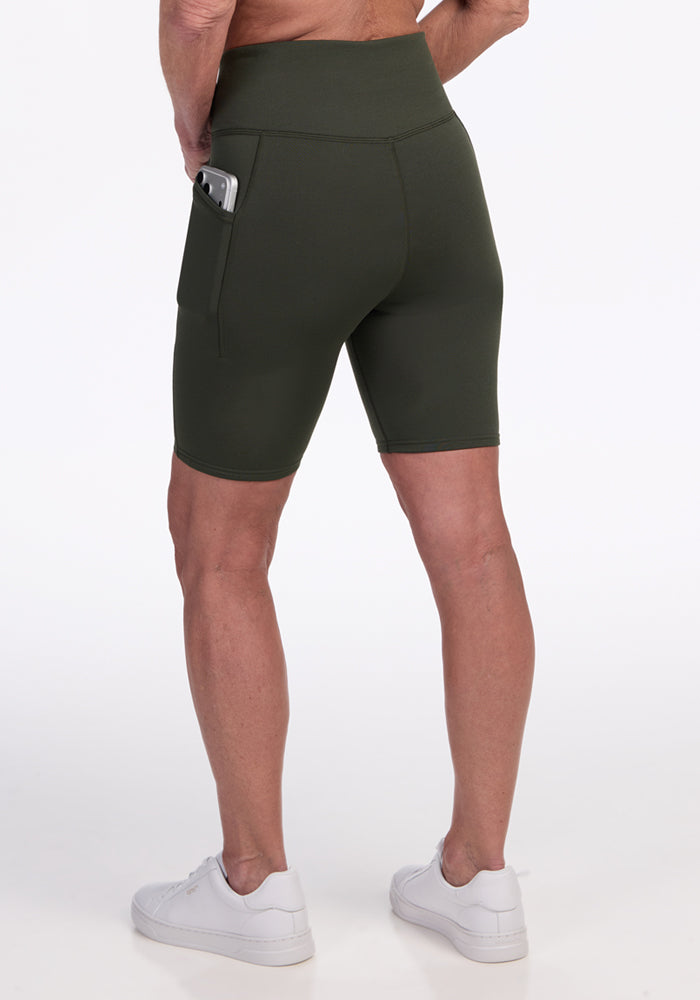 A person seen from behind wears Woolx Merino Flex® Dani Bike Shorts in Everleaf, featuring high-waisted, moisture-wicking fabric and a smartphone in the side pocket, paired with white sneakers against a plain white background.