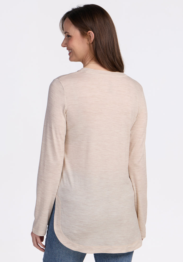 A woman with long brown hair, wearing the Woolx Hazel Tunic in Cream Heather and blue jeans, stands facing away, smiling over her shoulder against a plain white background.