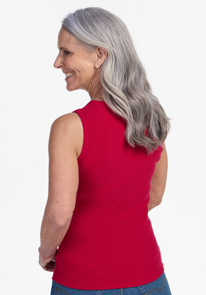 A smiling older woman with long gray hair wears the Woolx Raegan Ribbed Tank Top in Poppy Spark and blue jeans, standing with her back turned and head slightly to the side against a plain white background.