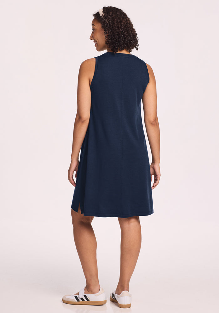 A woman with curly hair, wearing the Woolx Clara Dress in Deep Navy and white slip-on sneakers with black stripes, stands facing away from the camera against a plain light background.