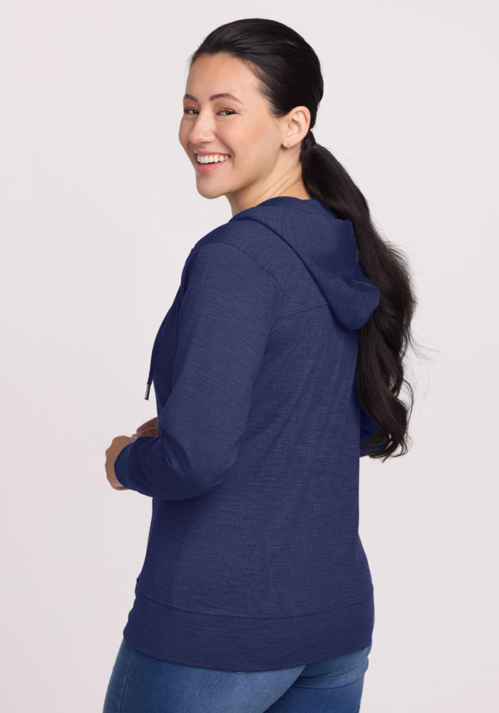Woman in Woolx Ryann Hoodie - Twilight Storm and jeans smiles, glancing over her shoulder against a light background.