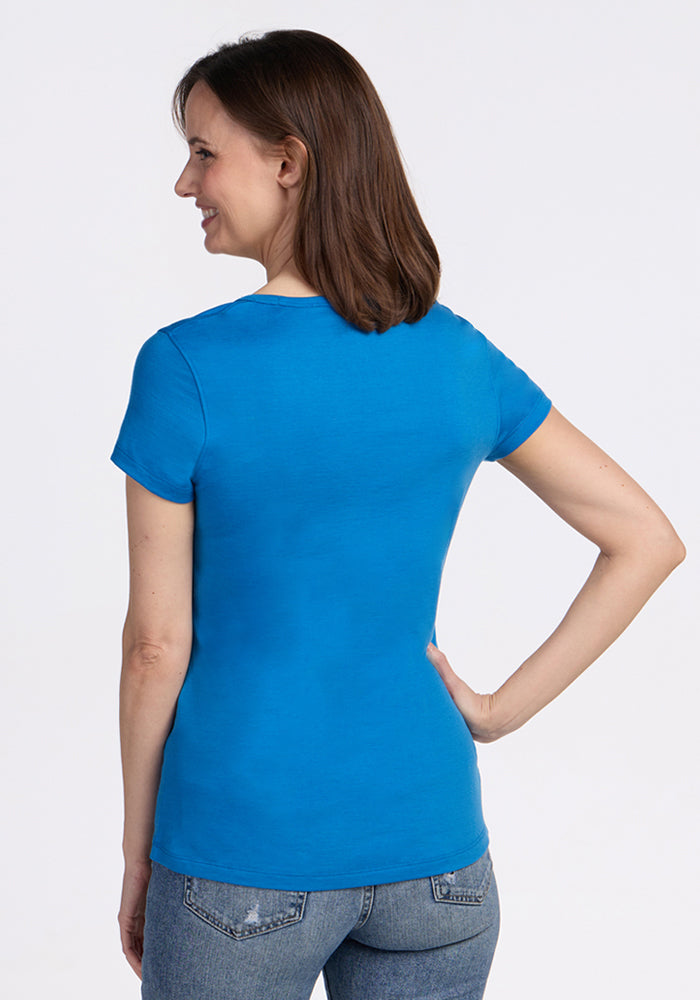 A woman with shoulder-length brown hair stands with her back to the camera, wearing the Woolx Addie Crew Neck - Wave Rider and blue jeans. One hand rests on her hip against a plain white background.