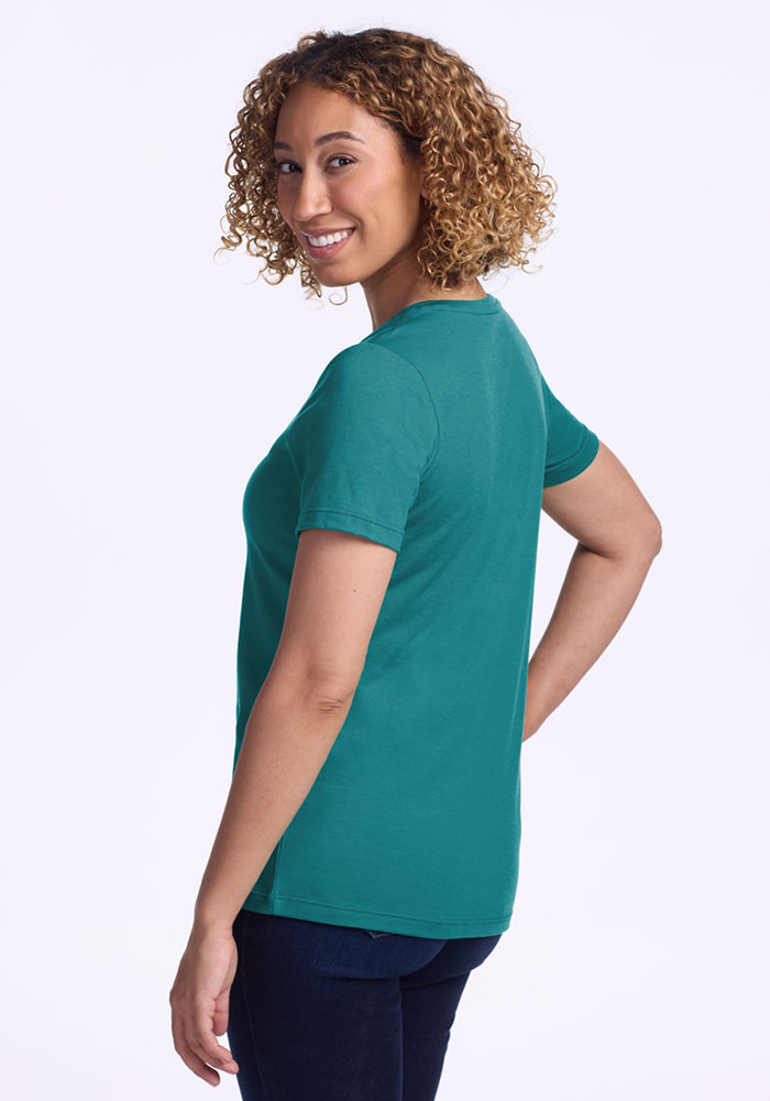 A woman with curly hair, wearing the Woolx Junie V Neck - Surfside and dark jeans, stands sideways and smiles at the camera against a plain white background.