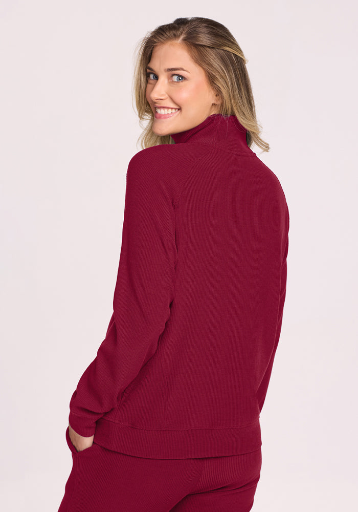 Woman in a Woolx Quinn Ribbed 1/4 Zip - Beet Red smiles over her shoulder, hands in pockets, on a plain background.