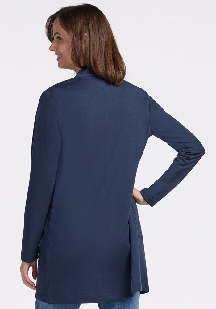 A woman with shoulder-length brown hair wears the Woolx Paisley Cardigan in Deep Navy and blue jeans, standing with her back to the camera as she looks over her shoulder and smiles.