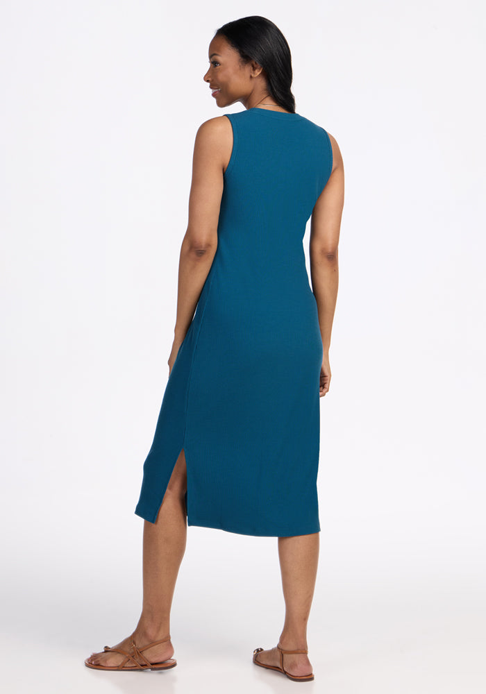 A woman stands facing away, wearing the Woolx Iris Ribbed Dress in Moroccan Blue with a side slit and brown flat sandals, against a plain white background.