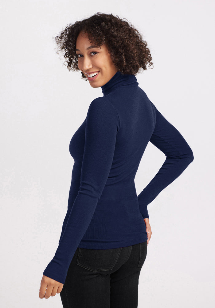 Smiling woman with curly hair in Woolx Sage Ribbed Turtleneck - Ocean Cavern and black jeans, glancing back.