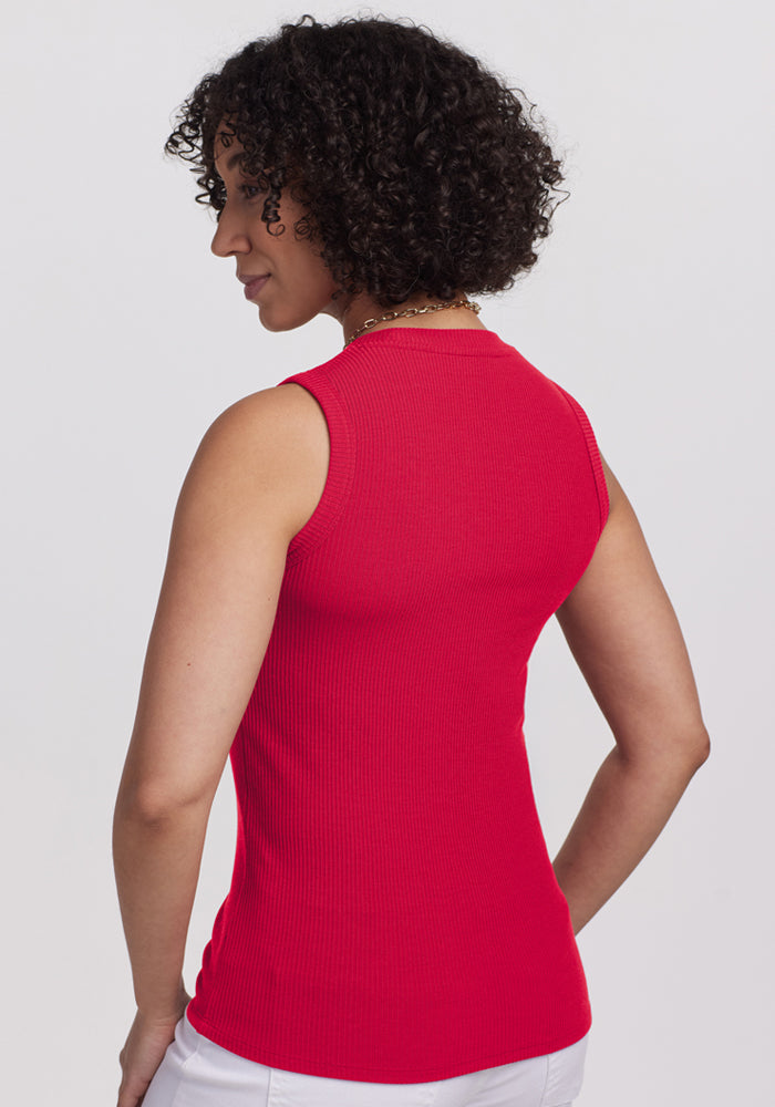A person with short curly hair, wearing the Woolx Sloane Ribbed Tank in Poppy Spark and white pants, stands with their back to the camera against a plain light background.