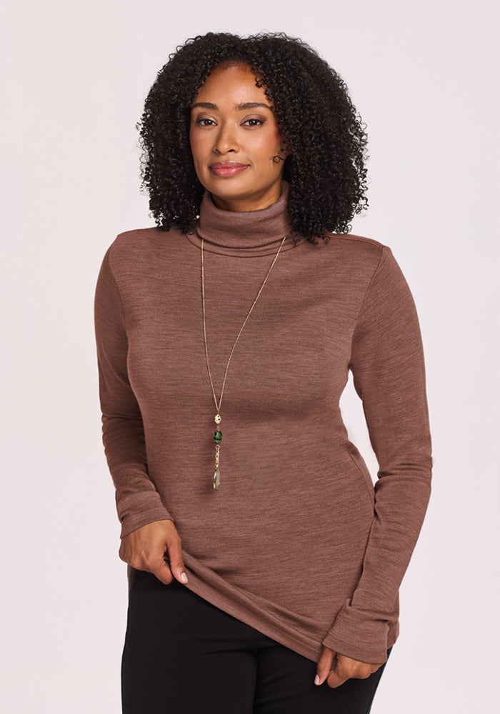 Woman with curly hair wears Woolx Peyton Turtleneck - Sable Heather and a long necklace, standing by a plain background.